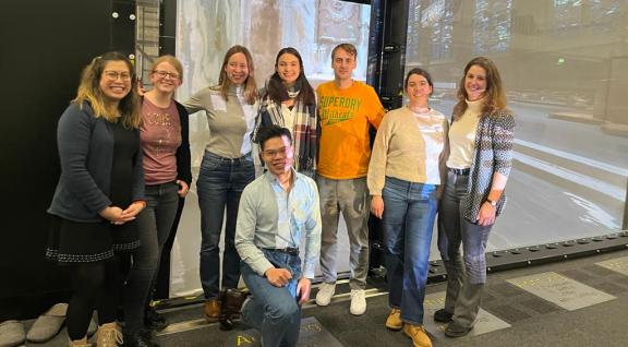 Six PhD students are lined up and smiling in front of the camera inside a university building