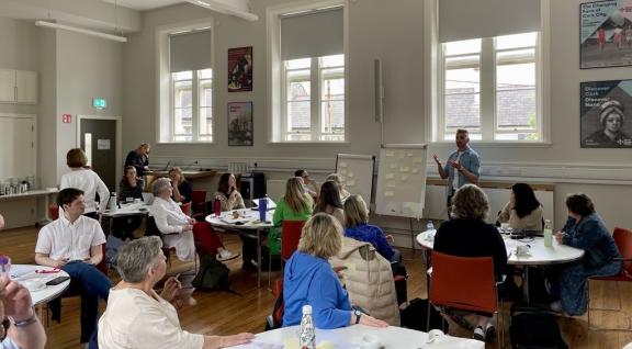 Someone is presenting facing a room full of people on separate round tables. They are all watching the presenter standing next to flipovers and in front of big windows.