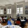 Someone is presenting facing a room full of people on separate round tables. They are all watching the presenter standing next to flipovers and in front of big windows.