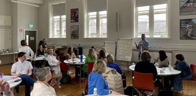 Someone is presenting facing a room full of people on separate round tables. They are all watching the presenter standing next to flipovers and in front of big windows.